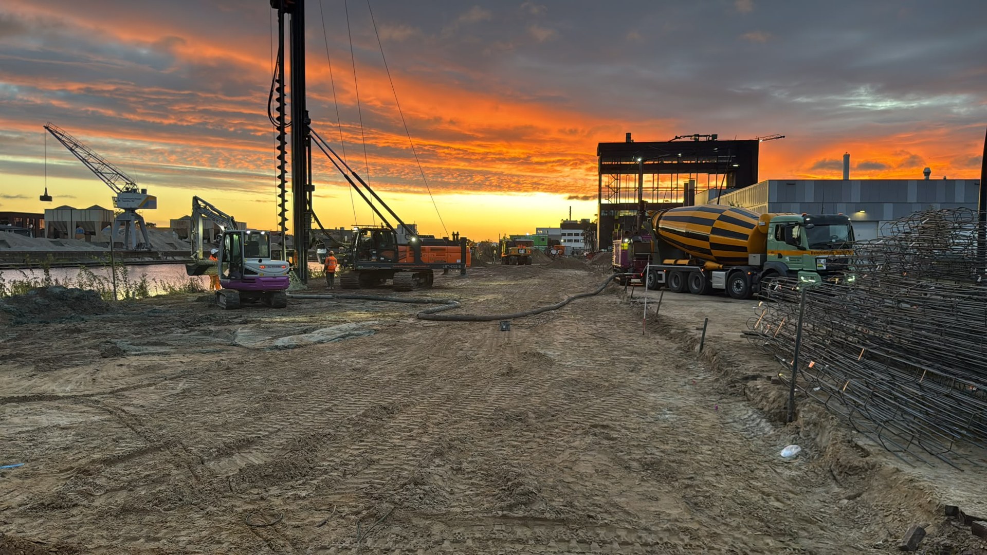 Boorinstallatie, betonmixer en wapening op de bouwplaats van Gebouw Q bij zonsopkomst.